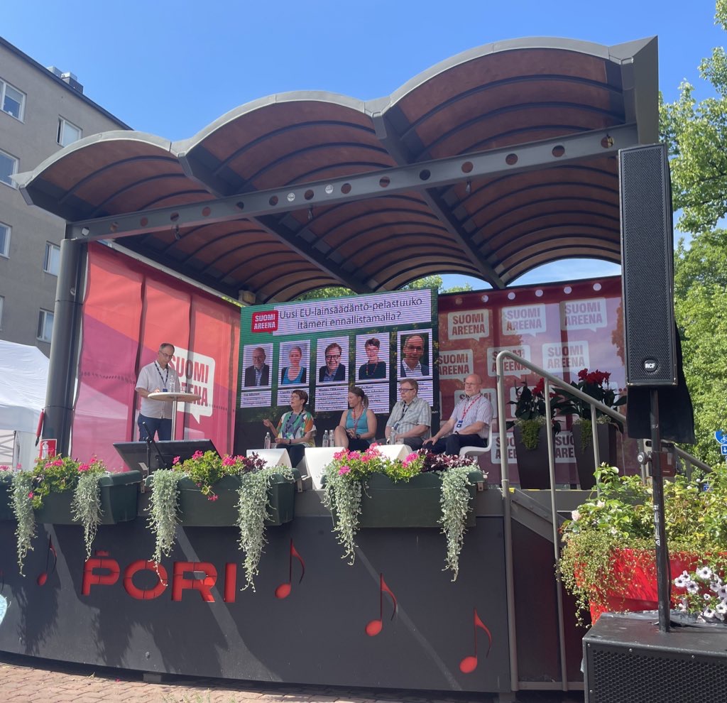 Valokuva ulkolavasta, jossa lukee Pori. Lavalla neljä panelistia istuu tuoleissa, yksi henkilö seisoo korkean pöydän luona. Lavan reunusta koristavat kukkaistutukset, taivas on sininen.
